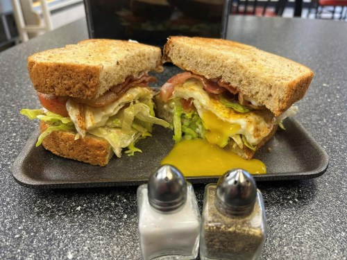 A close-up of a sandwich with lettuce, tomato, bacon, and a runny egg, served with salt and pepper shakers on a black tray.