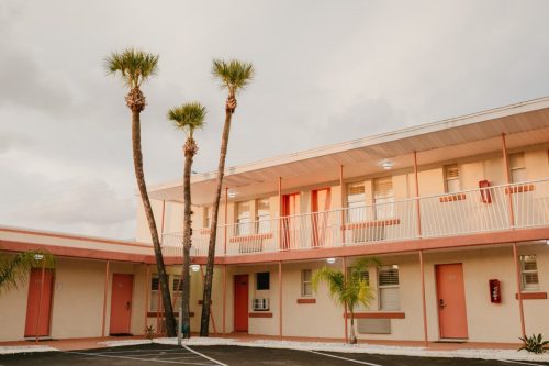 A pastel-colored motel with palm trees, featuring two levels and red doors under a cloudy sky.