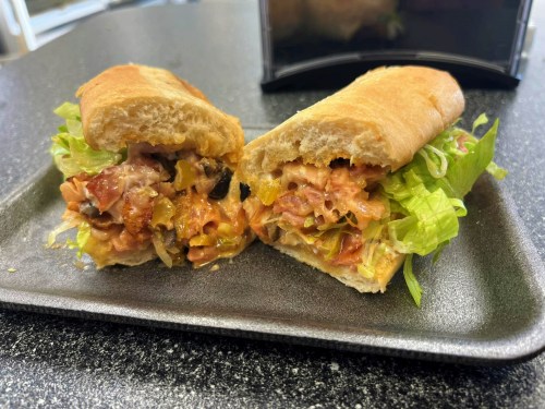 A sandwich cut in half, filled with lettuce, tomatoes, olives, and a creamy spread, served on a tray.
