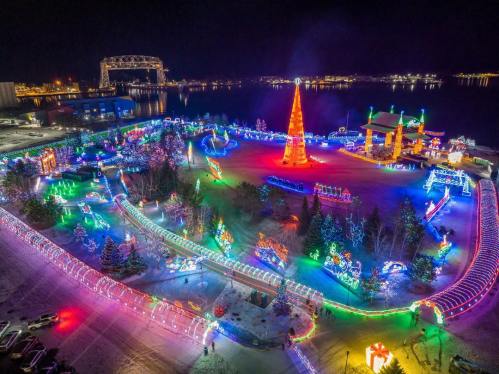 Aerial view of a vibrant holiday light display with colorful decorations and a large illuminated tree by a waterfront.
