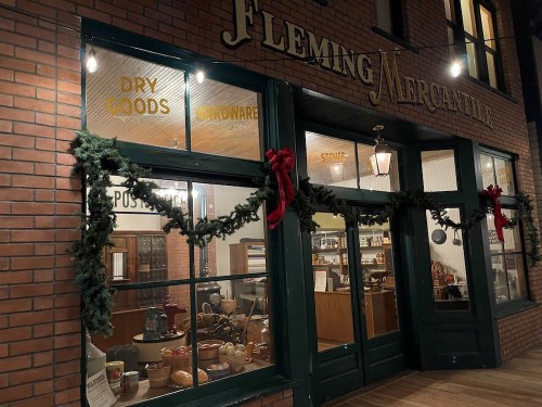 A festive storefront of Fleming Mercantile, decorated with garlands and bows, showcasing dry goods and hardware.