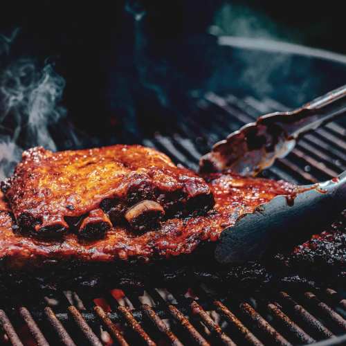 Close-up of juicy, glazed ribs sizzling on a grill, with smoke rising and tongs ready to serve.