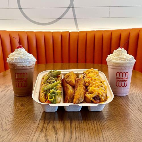 A tray with three loaded hot dogs and crispy potato wedges, flanked by two milkshakes topped with whipped cream and cherries.