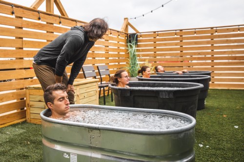 A group of people relaxes in metal tubs filled with water on a rooftop, surrounded by wooden fencing and greenery.