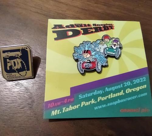 A colorful flyer for the Adult Soapbox Derby in Portland, Oregon, alongside two themed pins.