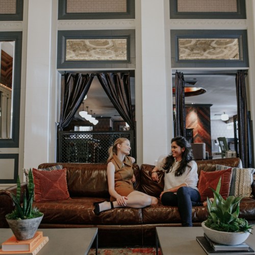 Two women sit on a brown leather couch in a stylish, well-lit lounge with plants and modern decor.