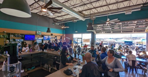 A busy café interior with patrons socializing, a bar area, and large windows showing an outdoor scene.