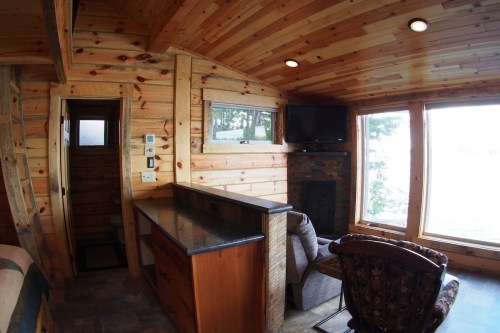 Cozy wooden interior of a cabin with a kitchen area, fireplace, and large windows overlooking a scenic view.