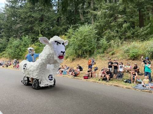 A person in a sheep costume on wheels parades past a crowd of spectators in a forested area.