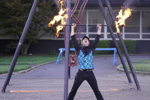 A performer in a black outfit and blue vest lifts a flaming staff, showcasing a fire act in a playground setting.