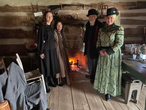 Four people in historical costumes stand by a fireplace in a rustic cabin, surrounded by wooden beams and decor.