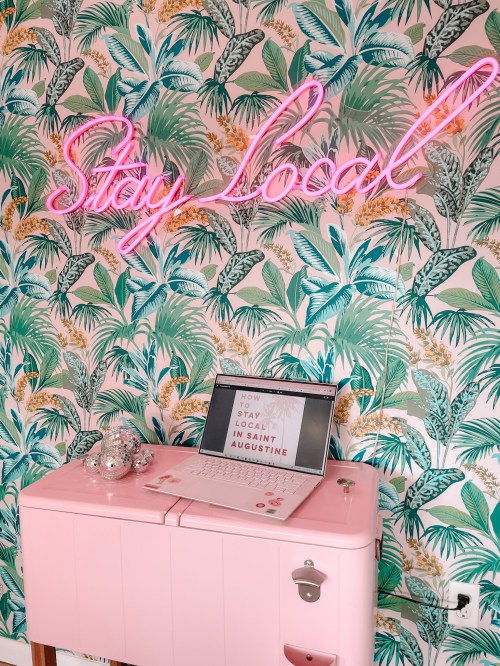 A pink desk with a laptop displaying "Stay Local" on a tropical leaf wallpaper background and a neon sign above.