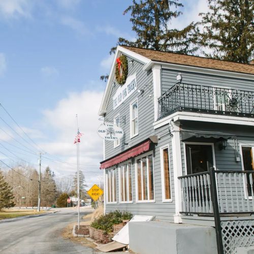 A charming gray building with a festive wreath, located at a roadside intersection, featuring a sign for "Old Oak Tavern."