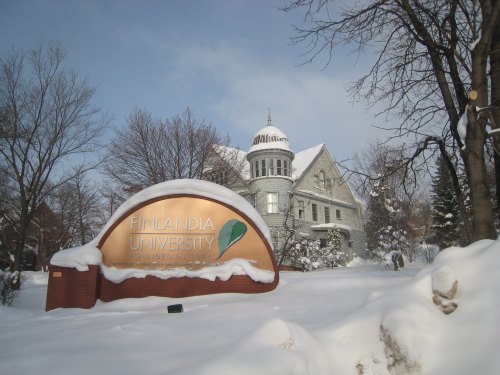 Snow-covered landscape featuring the Finlandia University sign and a historic building in the background.