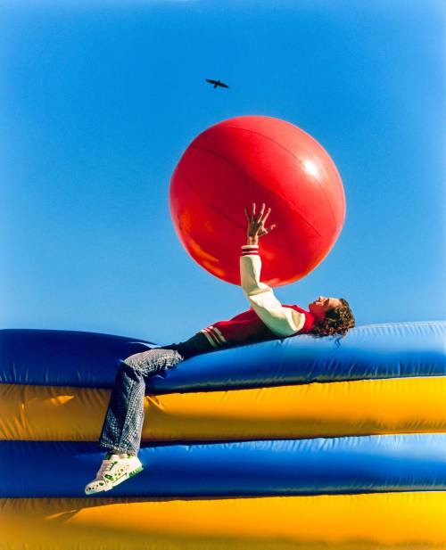 A person in a red and white jacket balances a large red ball while lying on a colorful inflatable surface under a blue sky.