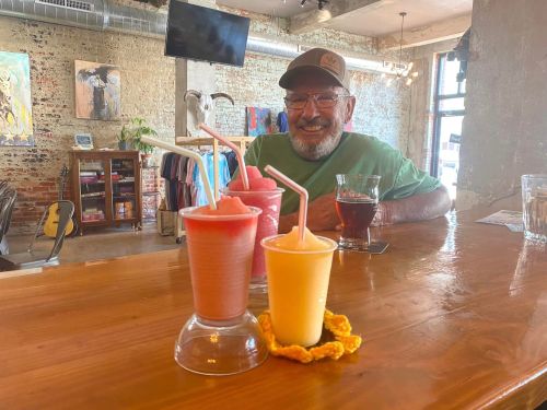 A smiling man sits at a bar with colorful frozen drinks in front of him, surrounded by a rustic interior.