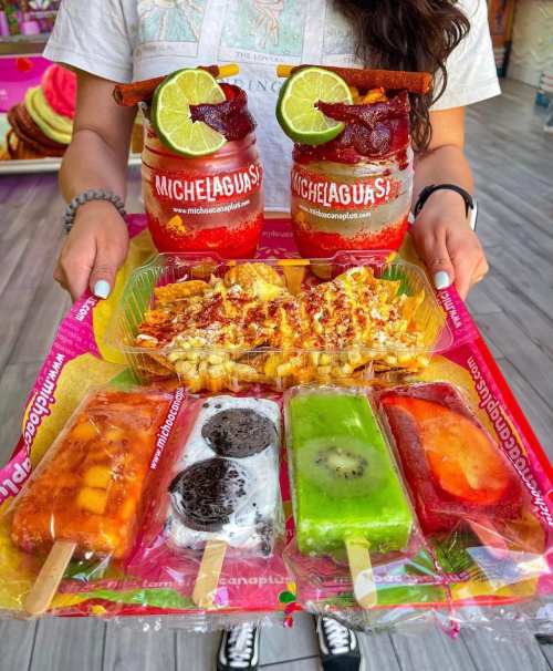 A person holds a colorful tray with various paletas, corn, and two drinks garnished with lime and bacon.