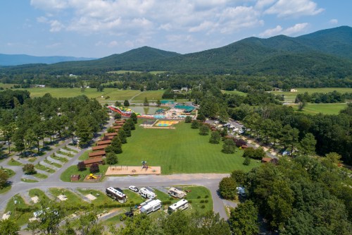 Aerial view of a campground with RVs, playground, and pool, surrounded by green fields and mountains in the background.