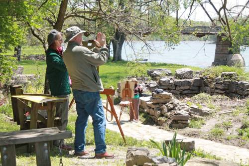 Two people outdoors by a river, one taking a photo while the other stands near a stone structure and a wooden table.