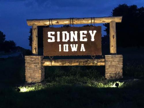 A wooden sign reading "SIDNEY IOWA" illuminated at night, surrounded by grass and trees.