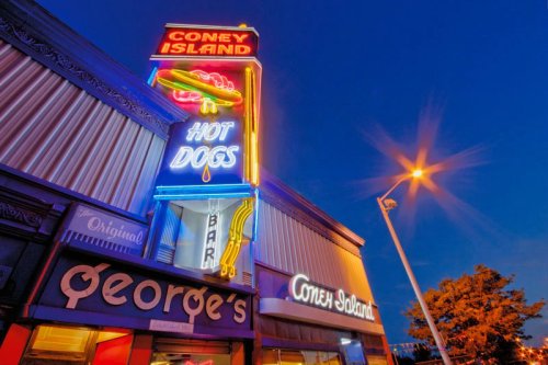 Neon sign for Coney Island hot dogs at dusk, featuring colorful lights and the name "George's" on the storefront.