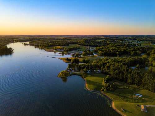 Aerial view of a serene lake surrounded by lush greenery and rolling hills at sunset.