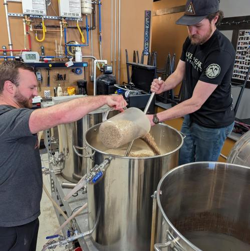 Two men are brewing beer, pouring ingredients into a large stainless steel kettle in a brewery setting.
