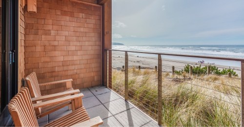 A balcony with wooden chairs overlooks a sandy beach and ocean under a clear sky.