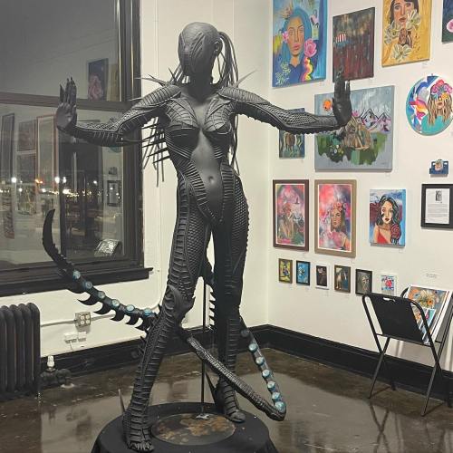 A striking black sculpture of a humanoid figure with intricate details, posed in an art gallery surrounded by colorful paintings.