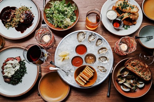A table spread with various dishes, including oysters, salads, and entrees, surrounded by drinks and candles.
