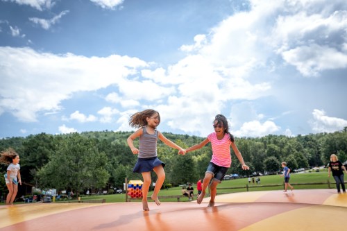 Two children joyfully jump on a colorful inflatable surface under a blue sky with fluffy clouds.