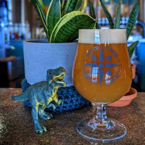 A glass of beer with foam sits on a counter next to a toy dinosaur and a potted plant.