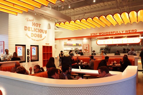 A vibrant restaurant interior with people dining, featuring bright orange decor and a menu highlighting hot dogs.
