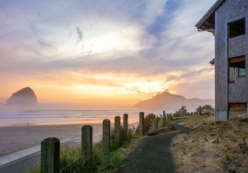 A serene beach scene at sunset, featuring a rocky outcrop and a wooden building beside a sandy path.