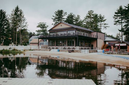 A modern lakeside building with large windows, surrounded by trees and reflecting in the water.