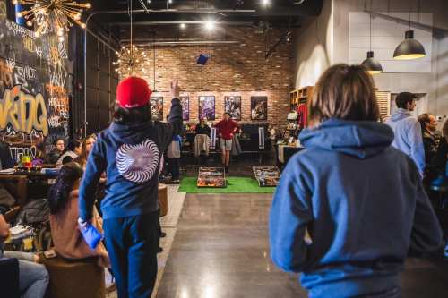 Two people in hoodies throw blue objects in a lively café with brick walls and artwork, while others socialize.