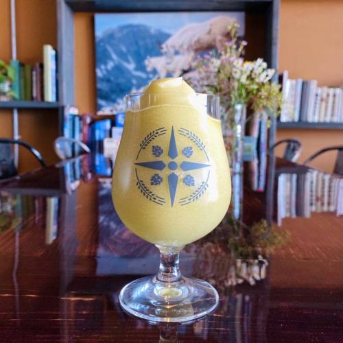 A frosty yellow drink in a decorative glass, placed on a wooden table with books and flowers in the background.