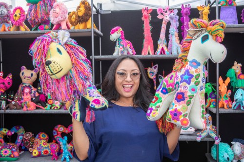 A woman smiles while holding colorful stuffed lion and unicorn toys, surrounded by vibrant plush animals on display.