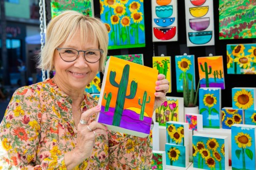 A smiling woman holds a colorful painting of cacti, surrounded by vibrant artwork featuring sunflowers and other designs.