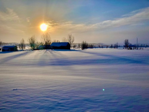 A serene winter landscape with a setting sun, snow-covered fields, and a silhouette of a barn and trees.