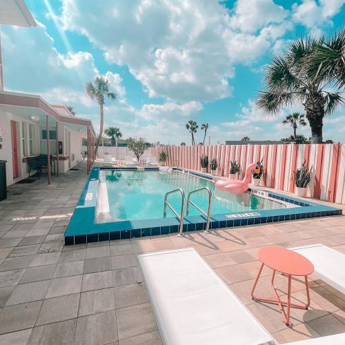 A sunny pool area with a pink flamingo float, lounge chairs, and palm trees under a blue sky with fluffy clouds.