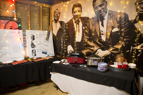 A festive setup with food, drinks, and a backdrop of vintage black-and-white portraits, illuminated by string lights.