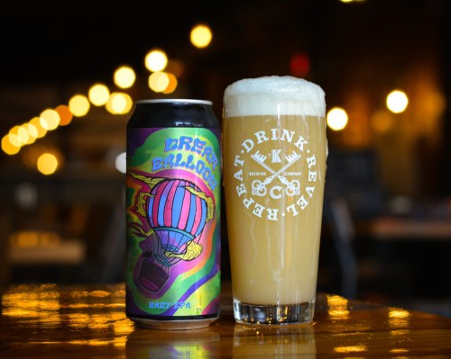A colorful beer can next to a glass of hazy beer, set against a blurred background with warm lights.