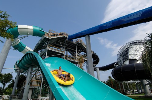 Two people ride a yellow inflatable tube down a water slide at a water park, with large slides in the background.
