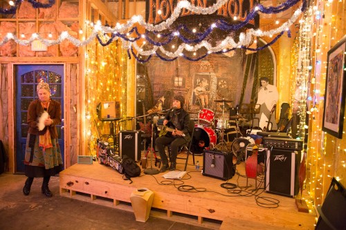 A cozy music venue with a band setting up, decorated with lights and festive decor, and a performer on stage.