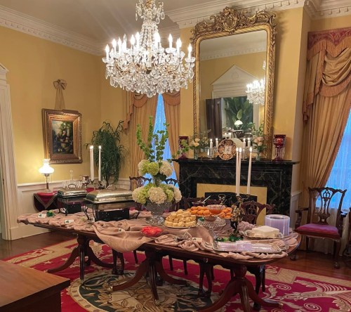 Elegant dining room with a lavish buffet table, chandelier, and ornate decor, featuring food and floral arrangements.