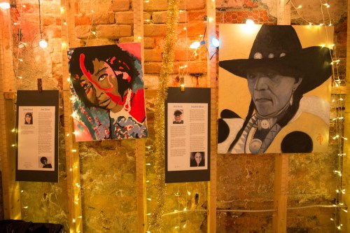 Three art pieces displayed on a wall, illuminated by string lights, featuring portraits and accompanying text descriptions.