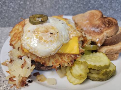 A plate with a crispy hash brown topped with a fried egg and cheese, served with pickles and toasted bread.
