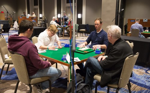 Four players engaged in a card game at a tournament, with green tables and spectators in the background.