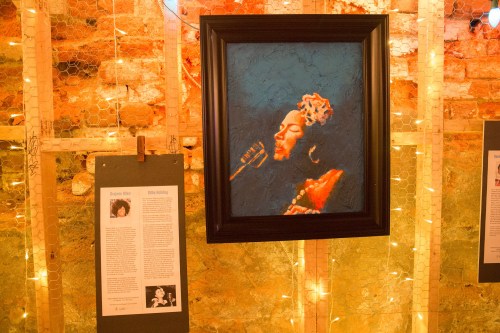 A framed painting of a woman singing, displayed on a wall with soft lighting and informational panels below.